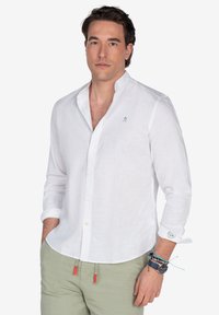 Camisa de lino blanca de manga larga con collar mandarín, adornada con bordados verde claro. Combinada con pantalones verde claro y pulseras en capas.