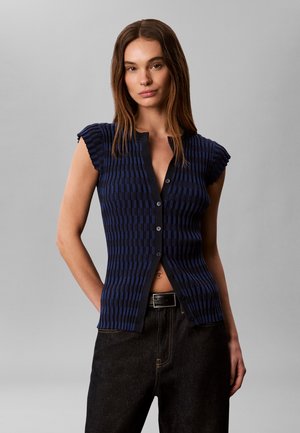 Femme portant un top texturé bleu marine à manches courtes et boutonné, et un jean noir avec une ceinture noire, debout devant un fond gris.