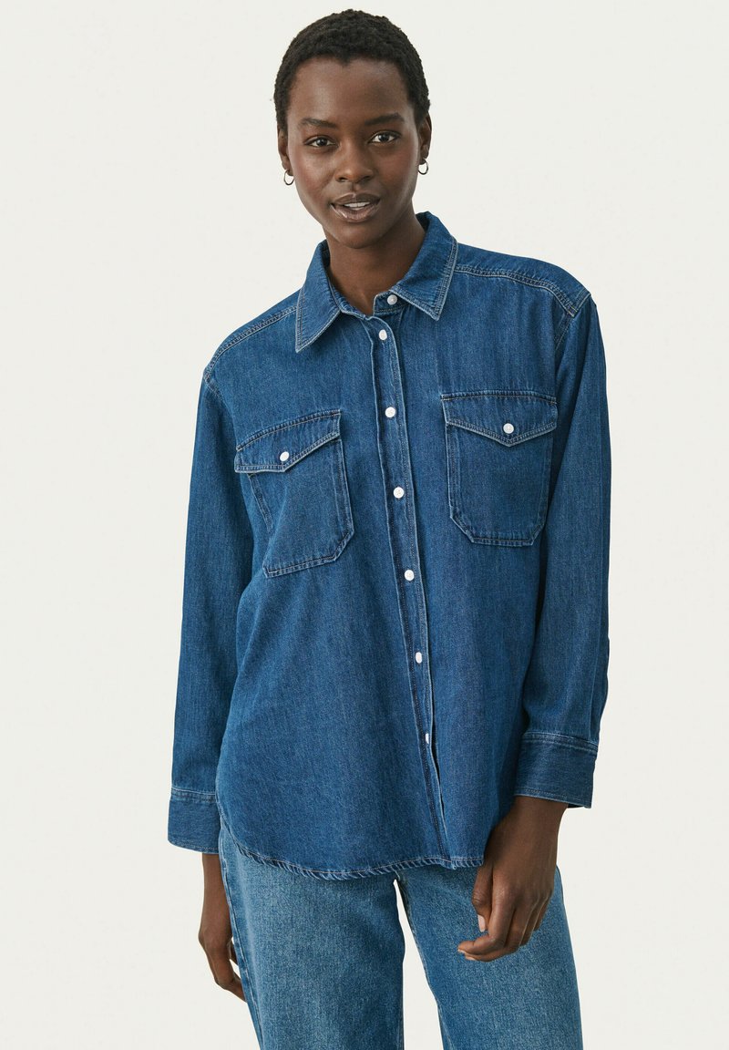 Camisa de denim con mangas largas, dos bolsillos en el pecho, cierre delantero con botones y dobladillo curvado en lavado azul oscuro. Textura suave, diseño clásico.