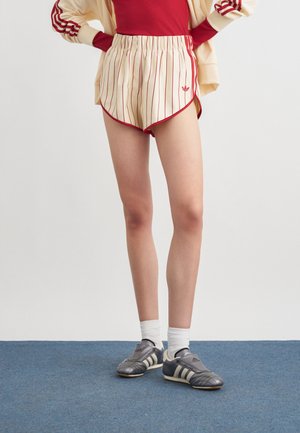 Persona che indossa pantaloncini crema con strisce verticali rosse, una maglietta rossa, calzini bianchi e scarpe Adidas nere e grigie, in piedi su un tappeto blu.