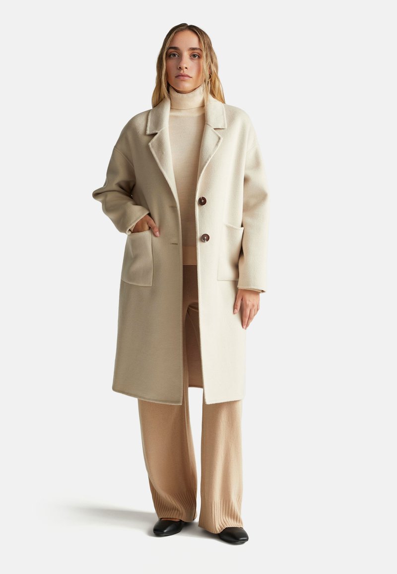 Cappotto in misto lana beige con colletto a punta, due tasche frontali e bottoni marroni. Indossato sopra un dolcevita beige coordinato e pantaloni.