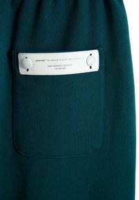 Tejido en tono teal que aparece en un diseño de pantalones de chándal con un bolsillo de etiqueta blanca. La etiqueta incluye el texto "MWM x mod wave movement" y logotipos.