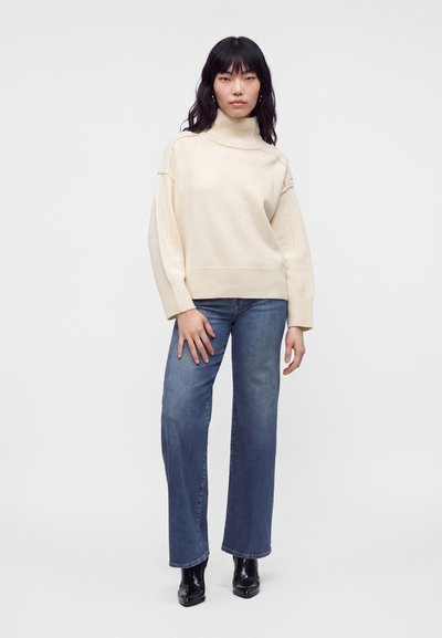 Femme aux longs cheveux foncés portant un pull à col roulé crème, un jean bleu à jambes larges et des bottines noires, debout devant un fond uni.