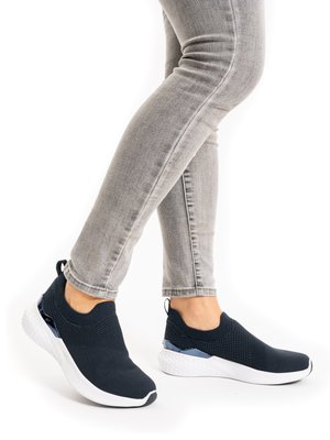Feet wearing black slip-on knit sneakers with white soles, paired with light gray fitted jeans against a white background.