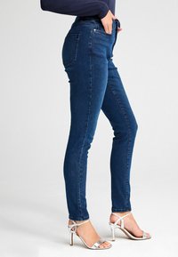 Uske mršave traperice u tamnoplavom denimu, s visokim strukom, pet džepova i suptilnim detaljima šivanja. Uparene s srebrnim sandalama na petu.