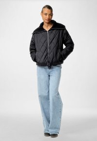 Must mustriga jope laia soonikkaga ja esi tõmbega, kombineeritud helesiniste laia säärega teksade ja tumedate kingadega. Tasane taust.