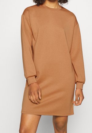 Nainen pukeutuneena pitkähihainen, kamelinvärinen college-mekko joustavilla resoreilla, seisoo yksinkertaisen vaalean taustan edessä.