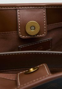Intérieur d'un sac en cuir marron avec une fermeture à pression magnétique dorée. Présente une texture lisse et des coutures contrastantes. Comprend une étiquette de marque.