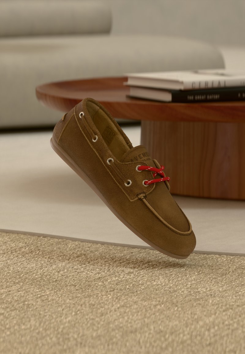 Καφέ μοκασίνι από σουέντ με κόκκινα κορδόνια που αιωρείται πάνω από μια υφή μπεζ χαλί, ένα στρογγυλό ξύλινο τραπεζάκι καφέ και στοιβαγμένα βιβλία στο φόντο.