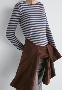 Person wearing a long-sleeve blue and brown striped shirt with a brown jacket tied around the waist, standing against a plain wall.