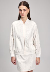 Giubbotto bomber bianco realizzato in tessuto testurizzato con dettagli a occhio e chiusura frontale con zip. Abbinato a una gonna coordinata, entrambi presentano schemi simili.