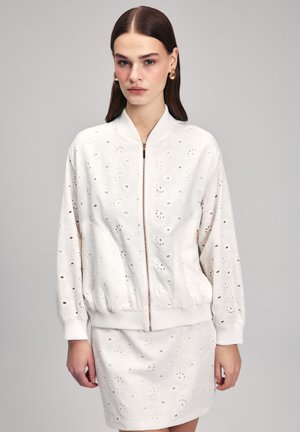 Giubbotto bomber bianco realizzato in tessuto testurizzato con dettagli a occhio e chiusura frontale con zip. Abbinato a una gonna coordinata, entrambi presentano schemi simili.