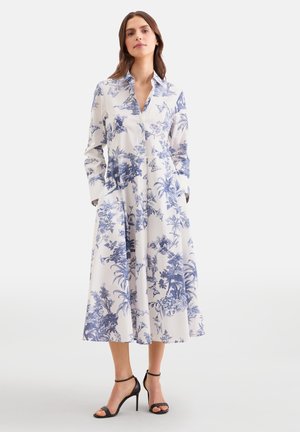 Frau trägt ein weißes Midikleid im Hemdstil mit blauen Blumen- und Schmetterlingsmotiven, langen Ärmeln und schwarzen Sandalen mit hohem Absatz.