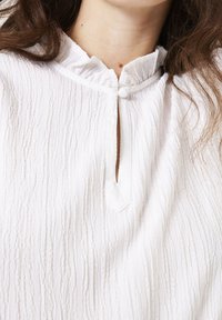Witte textuurblouse met een staande kraag, een gedeeltelijke knoopsluiting en een subtiel verticaal streeppatroon. De stof lijkt lichtgewicht en luchtig.