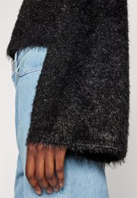 Main à la peau foncée reposant sur le côté, portant un pull noir duveteux et un jean bleu clair, gros plan montrant la texture du pull et les détails du denim.