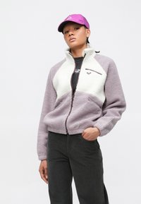 Jeune personne portant une veste polaire violette et blanche, un pantalon noir et une casquette violette, debout avec une main dans la poche.