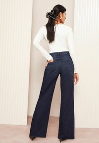 Frau mit dunklem Haar, das von einem schwarzen Haargummi zusammengehalten wird, trägt ein weißes langärmliges Oberteil und dunkle Jeans mit weitem Bein, steht mit einer Hand in der Gesäßtasche.