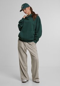 Oversized grøn hættetrøje med fronlomme, parret med lysegrå, løstsiddende bukser og grå slip-on sko. Grøn baseballcap med logo.
