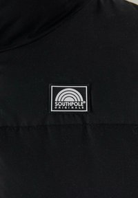 Sort stof med en glat tekstur, der har et rektangulært mærke med teksten "SOUTHPole ORIGINALS" og et buet regnbue-logo.