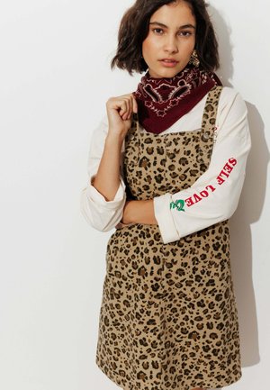 Femme aux cheveux courts et foncés portant une robe salopette à imprimé léopard, une chemise blanche à manches longues avec un texte rouge sur la manche, et un foulard rouge foncé à motifs autour du cou.
