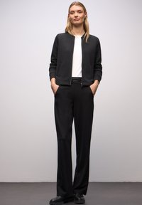 Zwarte doorgestikte bomberjas met een gestructureerd patroon, gecombineerd met zwarte op maat gemaakte broek en zwarte schoenen. Witte shirt met ronde hals eronder.