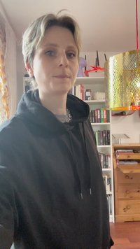 Felpa nera con cappuccio e lacci, dotata di una tasca frontale, indossata da una persona con capelli corti e chiari. Sospesa sopra c'è una lampada decorativa dorata.