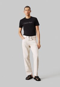 T-shirt en coton noir avec le logo "Calvin Klein", pantalon taille haute beige clair, et sandales noires à lanières, le tout sur un fond gris.