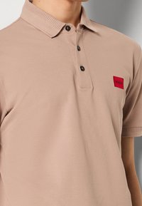 Jasnobeżowa koszulka polo z prążkowanym kołnierzykiem i krótkimi rękawami, z czerwoną prostokątną naszywką z logo "HUGO" na lewej piersi, z zapięciem na guziki.