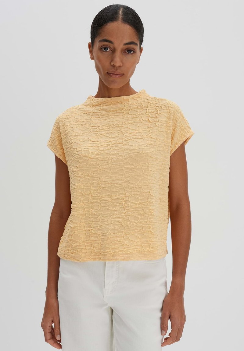 Kurze Ärmel, lässiges Oberteil aus strukturiertem, hellgelbem Stoff. Besitzt einen hohen Halsausschnitt und ein dezentes Wellenmuster. Kombiniert mit weißen Hosen.