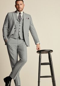 Traje de tres piezas gris claro con solapa muesca, corte ajustado y acentos marrones en contraste. Llevado con una camisa blanca, corbata de estampado y zapatos oscuros.