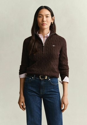 Frau mit langen dunklen Haaren, die einen braunen Zopfmuster-Viertel-Reißverschluss-Pullover über einem hellrosa Hemd und blauen Jeans mit schwarzem Gürtel trägt.