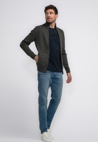 Stickad grå cardigan med hög krage, texturerade axlar, bärs över en marinblå t-shirt, i kombination med ljusblå jeans och vita sneakers.