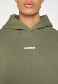 Personne portant un sweat à capuche vert olive avec le texte blanc "WRSTBHVR" sur la poitrine, visible des épaules aux lèvres sur un fond uni.