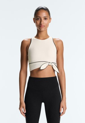 Kobieta z włosami upiętymi w kok, ubrana w bezrękawowy kremowy krótki top z czarną lamówką i wiązaniem z przodu, w połączeniu z wysokim stanem czarnych legginsów.