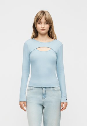 Žena oblečená v svetlomodrom tričku s dlhým rukávom a horizontálnym výrezom na hrudi, v svetlých rifliach, stojaca na bielom pozadí.