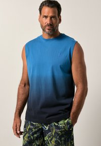 Man i ärmlös blå ombre skjorta och mörka shorts med grön lövtryck, stående med ena handen i fickan mot en enfärgad bakgrund.