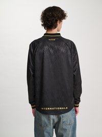 Person med krøllete hår iført en svart langermet humming sports shirt med gul "INTERNATIONALS"-tekst, kombinert med blå denim jeans, sett bakfra.