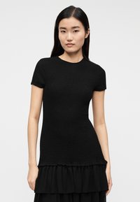 Νεαρή γυναίκα με ίσια σκούρα μαλλιά, που φοράει εφαρμοστό μαύρο τοπ με υφή και κοντά μανίκια, συνδυασμένο με μαύρη φούστα με βολάν.