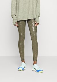 Oliwkowe legginsy z metalicznymi wzorami liści, w połączeniu z jasnobłękitnymi i białymi trampkami. Całość uzupełnia przewiewny szary top w dużym rozmiarze.