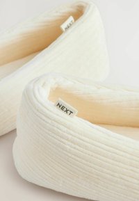 Chaussons de couleur crème en tissu peluche côtelé avec un extérieur doux et texturé et une étiquette visible portant la mention "NEXT".