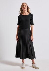 Svart klänning med korta ärmar, plisserad kjol och åtsittande liv. Bärs med lätta sneakers för en avslappnad look. Slät textur, enfärgad.