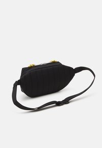 Bolsa de cintura negra acolchada con correa ajustable y tiradores de cremallera plateados con cordones amarillos, contra un fondo blanco liso.