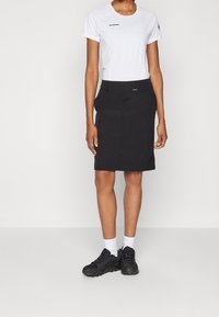 Camisa blanca de manga corta con un logo negro combinada con una falda negra hasta la rodilla; modelo lleva zapatillas negras y calcetines blancos.