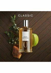 Flaska med David Beckham Classic doft. Klar glas med cylindrisk form, guldvätska inuti, silverlock och vit etikett. Ackompanjerad av gröna blad och en lime.