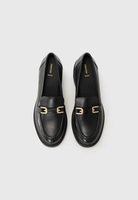 Mocassins en cuir noir avec des accents en métal doré, présentant un bout arrondi et une surface texturée. Design minimaliste adapté à diverses occasions.