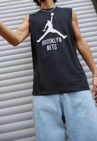 Persoon in een mouwloos zwart Brooklyn Nets shirt met een wit logo van een basketbalspeler en lichtblauwe jeans, staand voor een gesloten achtergrond.