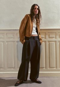 Jeune femme aux longs cheveux portant une veste en daim marron, un haut blanc, un pantalon large noir et des chaussures plates noires, debout à l'intérieur contre un mur à panneaux.