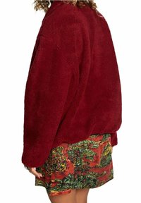 Röd oversized fleecejacka med en texturerad yta, tillsammans med en färgglad mönstrad kjol med växtlighet och livfulla färger.