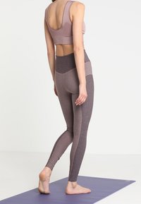 Jasnofioletowy stanik sportowy oraz legginsy z wysokim stanem, z teksturowanym wzorem w kontrastujących odcieniach fioletu i szarości, na macie do jogi.