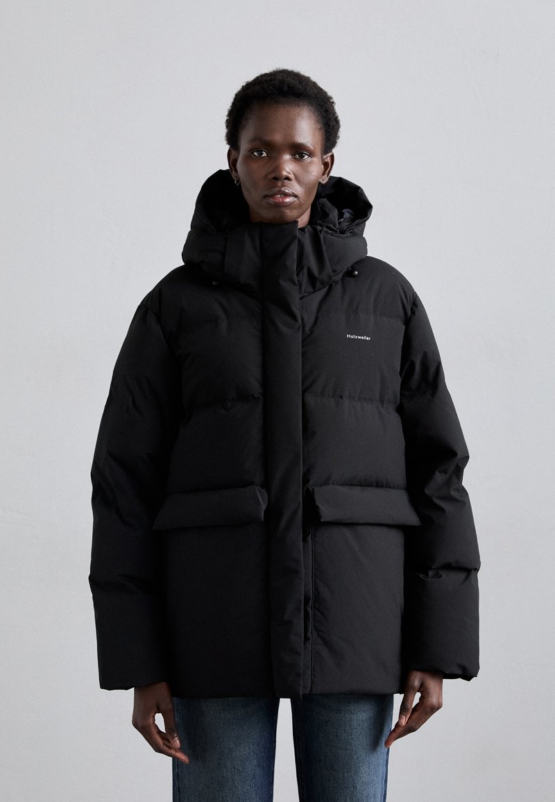 Black puffer jacket with a high collar, oversized fit, front pockets, and a hood. Smooth fabric with a matte finish. Brand logo on the chest.